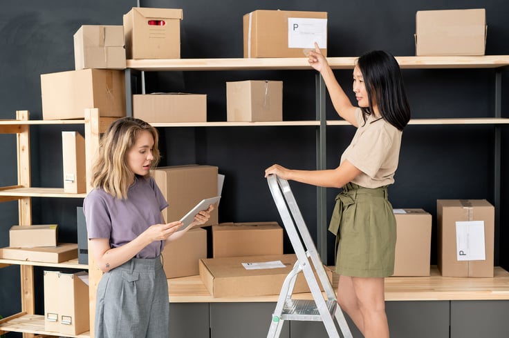 two-girls-working-in-store-room-by-large-shelf-wit-6RTRBLB